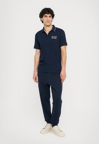 Marineblå polo med et gyldent logo, korte ærmer og kontrastkrave; kombineret med matchende marineblå joggers og lyse sneakers.
