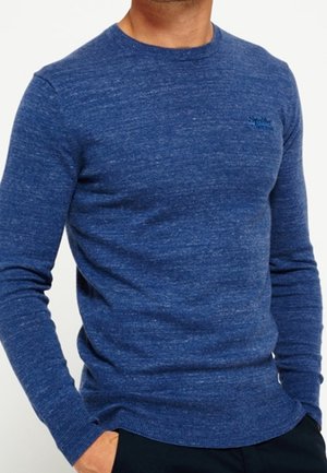 Mann trägt einen eng anliegenden blauen Pullover mit Rundhalsausschnitt, langen Ärmeln und dezent strukturierter Strickoptik, Hände in den Taschen.