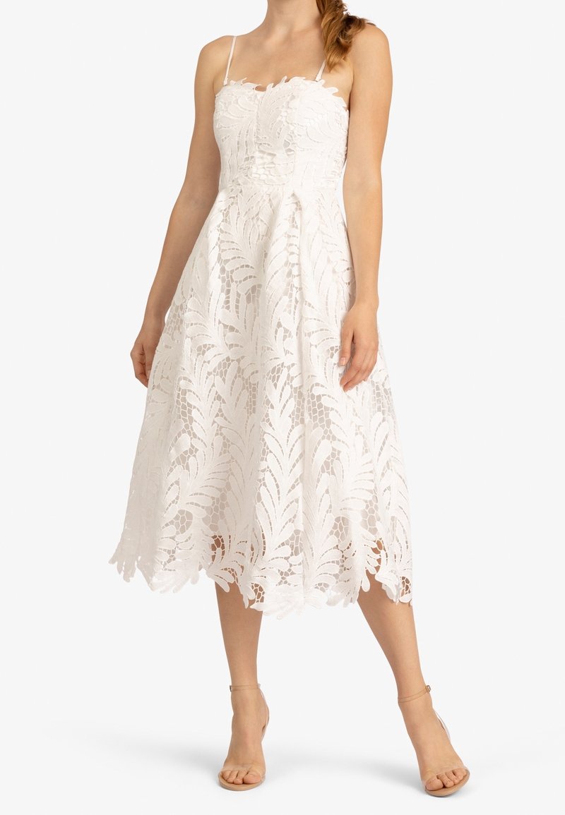 Femme portant une robe midi blanche sans manches en dentelle avec fines bretelles et des talons nude à bride cheville, debout devant un fond uni.