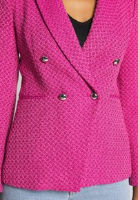 Femme portant un blazer texturé rose vif à double boutonnage avec des boutons argentés, par-dessus un haut noir et un jean bleu clair.