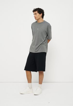 Νέος άνδρας στέκεται με τα χέρια πίσω από την πλάτη, φορώντας γκρι μπλουζάκι, μαύρα σορτς μέχρι το γόνατο, λευκές κάλτσες και λευκά αθλητικά παπούτσια, μπροστά από λευκό φόντο.