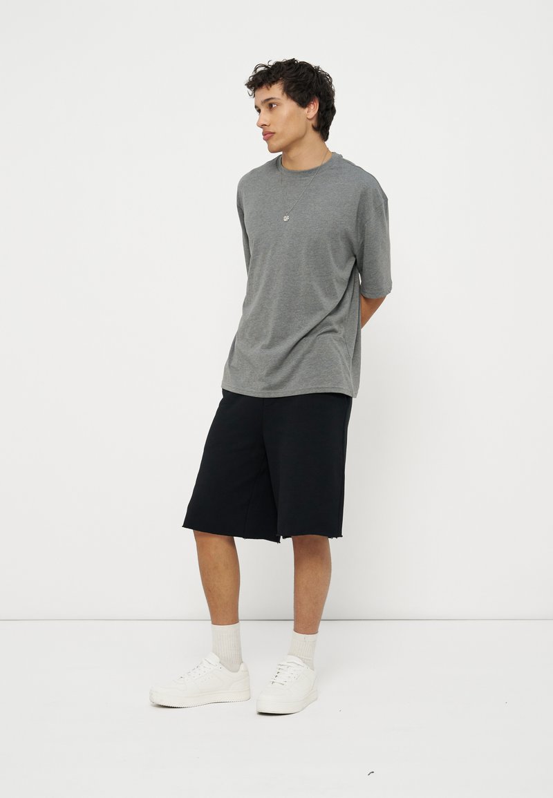 Νέος άνδρας στέκεται με τα χέρια πίσω από την πλάτη, φορώντας γκρι μπλουζάκι, μαύρα σορτς μέχρι το γόνατο, λευκές κάλτσες και λευκά αθλητικά παπούτσια, μπροστά από λευκό φόντο.