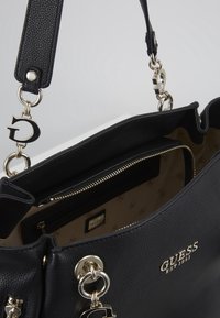 Zwarte gestructureerde Guess-handtas met gouden hardware, logo charms en beige voering met een ritszak en merkembleem.