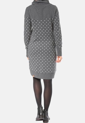 Vrouw die een grijze, lange mouwen, polkadot jurk met kap draagt, zwarte panty's en zwarte enkellaarzen, van achteren gezien.