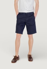 Pantaloni corti in cotone blu navy con frontale piatto, tasche laterali e chiusura a bottone. Indossati con una camicia bianca e scarpe stringate marroni.