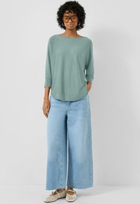 Kobieta z kręconymi włosami, nosząca duże bursztynowe okulary przeciwsłoneczne, luźną turkusową bluzkę, szerokie jasnoniebieskie dżinsy i plecione beżowe baleriny na tle jednolitym.