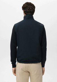 Zwarte vest met een hoge kraag en verschillende texturen; gevoerde schouderpanelen en zachte mouwen; gedragen over een paar beige broeken.