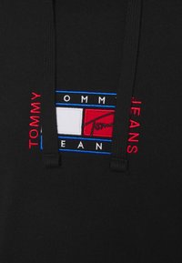 Sweat à capuche noir avec le logo Tommy Jeans présentant un drapeau rectangulaire rouge, blanc et bleu ainsi qu'un texte vertical rouge de chaque côté du logo.