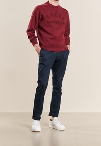 Person wearing a burgundy Steve McQueen sweatshirt, navy blue rolled-up pants, and white sneakers standing with hands in pockets.