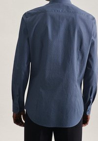 Man som bär en långärmad, mönstrad blå skjorta sedd bakifrån, tillsammans med mörka byxor mot en enkel bakgrund.