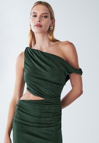 Emeraldgroene jurk met één schouder, met geplooide details, een uitgesneden middenstuk en een zachte textuur. Geaccentueerd door een gedraaid ontwerp en geplooide stof.