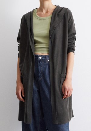 Personne portant un cardigan à capuche gris foncé par-dessus un t-shirt court vert clair et un jean bleu taille haute, sur un fond uni.