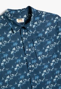 Camisa de botones de manga corta, color azul oscuro, con un patrón de palmeras y veleros. Presenta detalles en azul claro y un bolsillo tipo parche en el pecho.