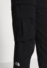 Svarta cargobyxor med flera fickor, tillverkade av slitstarkt tyg. Har sömnadsdetaljer och en logotypapplikation nära nedre ben.