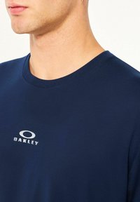 Navy blue cotton t-shirt with a round neckline and short sleeves, featuring a silver "OAKLEY" logo on the chest. Smooth texture.
