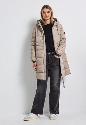 Beige Daunenjacke mit verstellbarem Kapuzenzug, Reißverschluss und gerippten Bündchen, kombiniert mit schwarzen weit geschnittenen Jeans und weißen Turnschuhen.