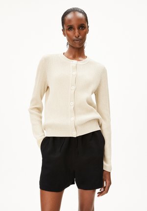 Beige ribbeltrui met een ronde hals, zes knopen, en lange mouwen, gecombineerd met zwarte shorts. Gelaagd, comfortabel breiwerk.