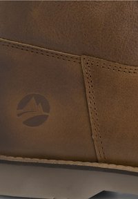 Gros plan d'une chaussure en cuir marron avec une couture détaillée et un logo de montagne en relief sur le côté au-dessus d'une semelle marron clair.