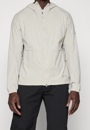 Person trägt eine hellbeige Kapuzenjacke mit Reißverschluss und elastischen Bündchen sowie schwarze Hosen, steht vor einem einfarbigen Hintergrund.