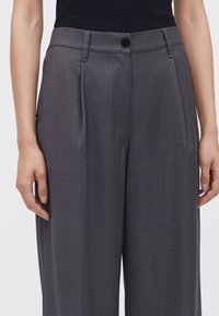 Taille et haut des jambes d'une personne portant un pantalon gris taille haute à jambes larges avec plis sur le devant et fermeture par bouton noir.