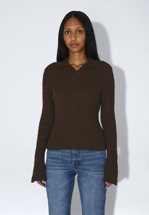 Donna con lunghi capelli neri che indossa un maglione marrone a coste a maniche lunghe e jeans blu, in piedi davanti a uno sfondo bianco semplice.