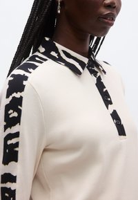 Krämfärgad pikétröja med svartvita mönstrade detaljer på axlarna; har krage med struktur och knapplugg.