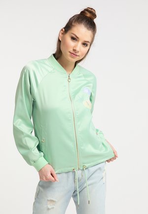 Giovane donna con i capelli legati indossa una giacca verde chiaro con cerniera e jeans azzurri strappati, posando con le mani nelle tasche su sfondo bianco.