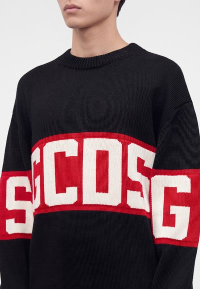 Personne portant un pull en maille noir avec une bande rouge et blanche à travers la poitrine et les manches, présentant de grandes lettres en gras.