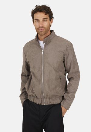 Mężczyzna z krótkimi kręconymi włosami, ubrany w jasnobrązową kurtkę na zamek na białą koszulę, z rękami w kieszeniach czarnych spodni.