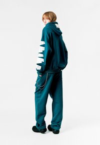Conjunto de sudadera y pantalones en color verde azulado con un diseño de zigzag blanco en las mangas, ajuste relajado y un detalle de cadena plateada en los pantalones. Tejido texturizado.