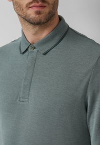 Gros plan d'un homme portant un polo vert texturé avec col boutonné et bords arrondis sur un fond neutre.