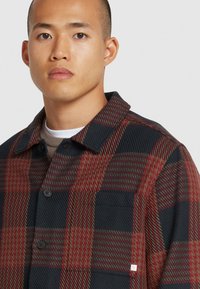 Camicia in flanella a quadri nei toni del blu navy scuro, bordeaux e verde oliva. Presenta un colletto, chiusura a bottoni e una tasca sul petto. Tessuto testurizzato.