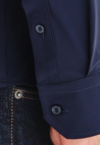 Granatowy mankiet koszuli z kontrastującym guzikiem; gładka, błyszcząca tkanina; kanciasty design; widoczne detale szwów; poniżej widoczna tkanina dżinsowa.