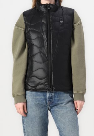 Personne portant un gilet matelassé noir sans manches par-dessus un sweat-shirt vert à manches longues et un jean bleu clair, debout devant un fond uni.