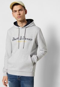 Grå hættetrøje med snor, logo i mørkeblå og gul, frontlomme, ribbede håndled og kant. Bæres med blå jeans, person står mod en hvid baggrund.