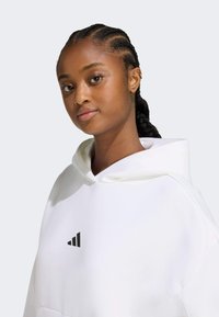 Biely mikina s veľkou kapucňou a čiernym logom Adidas na hrudi. Má jemnú textúru a voľný strih, rámovaný vrkôčkami.