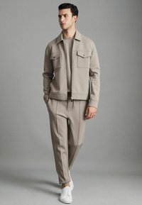 Jachetă zip-up de culoare gri deschis, cu două buzunare pe piept, asortată cu pantaloni corespunzători. Material texturat, croială lejeră, completată cu adidași albi.
