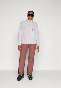 Άτομο που φορά σκούρα γυαλιά ηλίου, ανοιχτό μωβ φλις με φερμουάρ και λευκή τσέπη, ροζ παντελόνια και μαύρα δέσιμο παπούτσια, που στέκεται σε απλό φόντο.