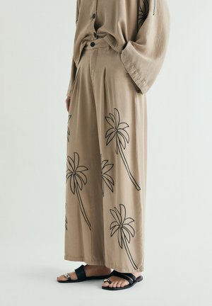 Pantalones anchos beige con contornos negros de palmeras, combinados con un top de manga larga a juego y sandalias negras de punta abierta.