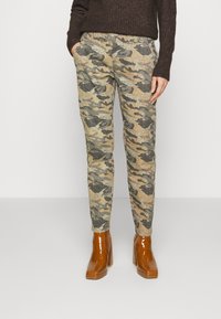 Calças com padrão camuflado em tons de verde e castanho suaves, apresentando uma cintura confortável e bolsos laterais, acompanhadas de botas de salto castanhas brilhantes.