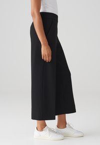 Pantalons larges noirs avec une texture lisse et une taille élastique, associés à des baskets blanches, montrés de c�ôté.