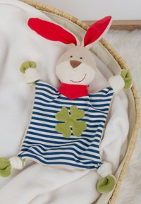 Soft bunny toy with red ears, blue striped body, green clover patch, and knotted limbs resting on a white blanket in a wicker basket.