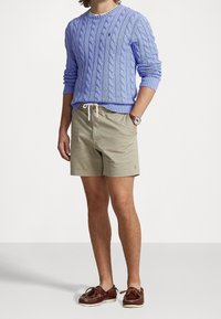 Man som bär en ljusblå kabelstickad tröja, beige dragkedjeshorts, brunt läderarmband och bruna läderbåtskor.