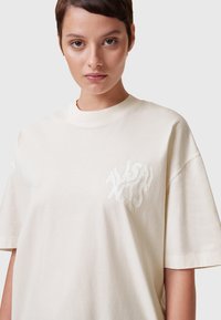Camiseta de algodón oversize color crema con cuello redondo y mangas cortas, que presenta un logo impreso en blanco texturizado en el pecho.