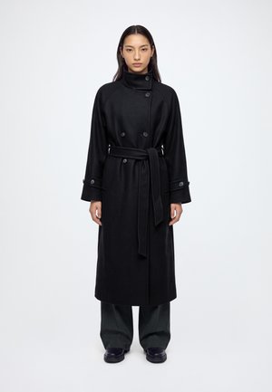 Femme debout portant un long manteau noir ceinturé sur un pantalon large foncé et des chaussures noires sur fond blanc uni.