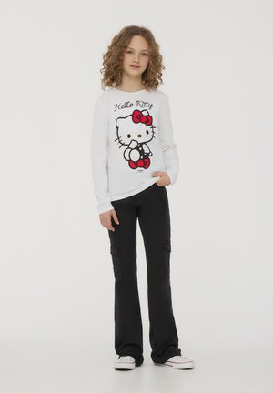 T-shirt bianco a maniche lunghe con grafica di Hello Kitty in nero e rosso, abbinato a pantaloni cargo neri; ragazza in piedi con le mani nelle tasche.