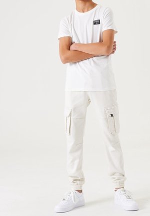 Person trägt weiße Cargo-Joggerhosen, weiße Sneakers und ein weißes kurzärmeliges T-Shirt mit kleinem Logo auf der Brust, die Arme verschränkt.