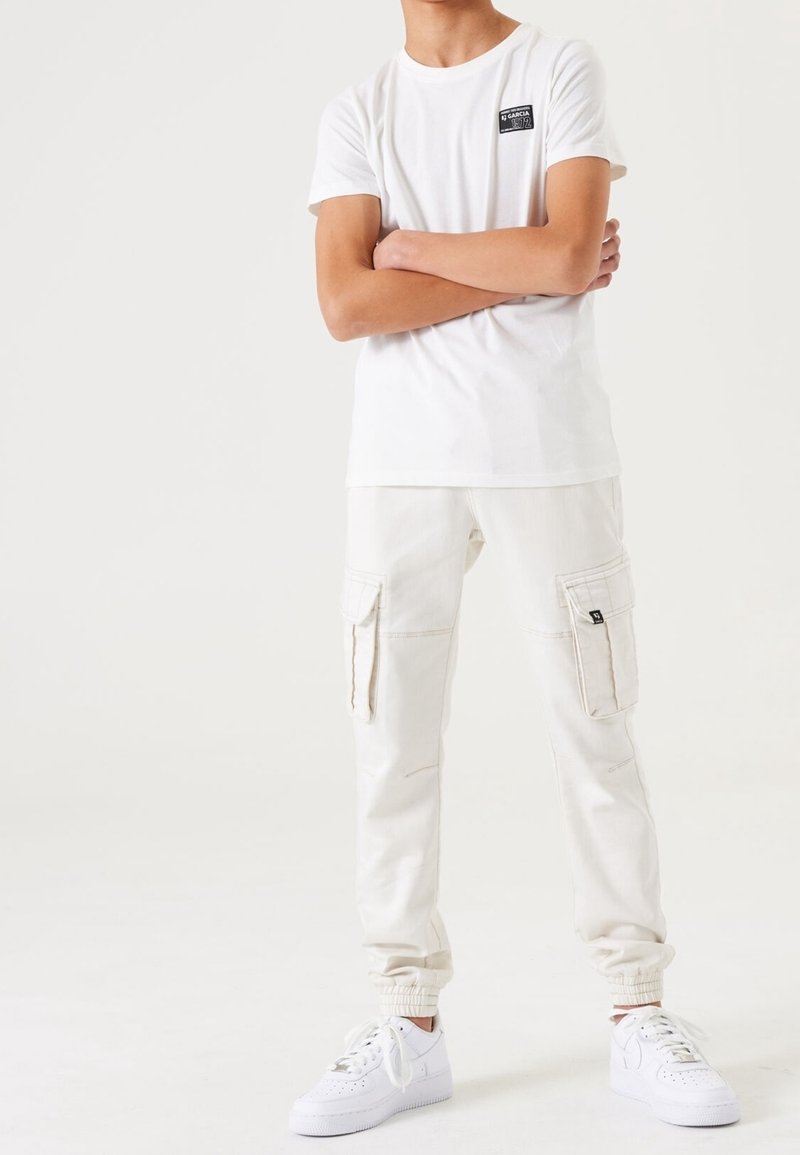 Personne portant un pantalon cargo jogger blanc, des baskets blanches et un T-shirt blanc à manches courtes avec un petit logo sur la poitrine, les bras croisés.