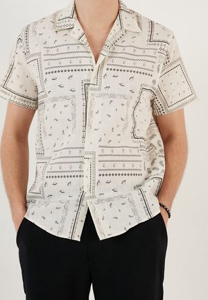 Mann trägt kurzärmliges Hemd mit Knöpfen, schwarzem und grauem Paisley- und geometrischem Muster, kombiniert mit schwarzer Hose und Armband.