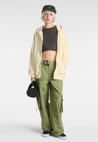 Gul hoodie, mörk kort överdel och olivgröna cargobyxor med fickor. Svart keps och rund svart väska. Sneakers fulländar outfiten.